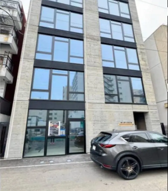 大きな窓が特徴のモダンなデザインのビルで、前には駐車場と車が写っている
                                                